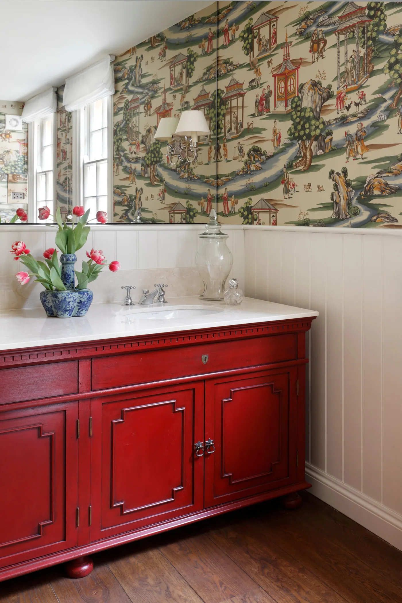 Red paneled bathroom vanity beneath scenic wallpaper, a pale counter and a large wall mirror.