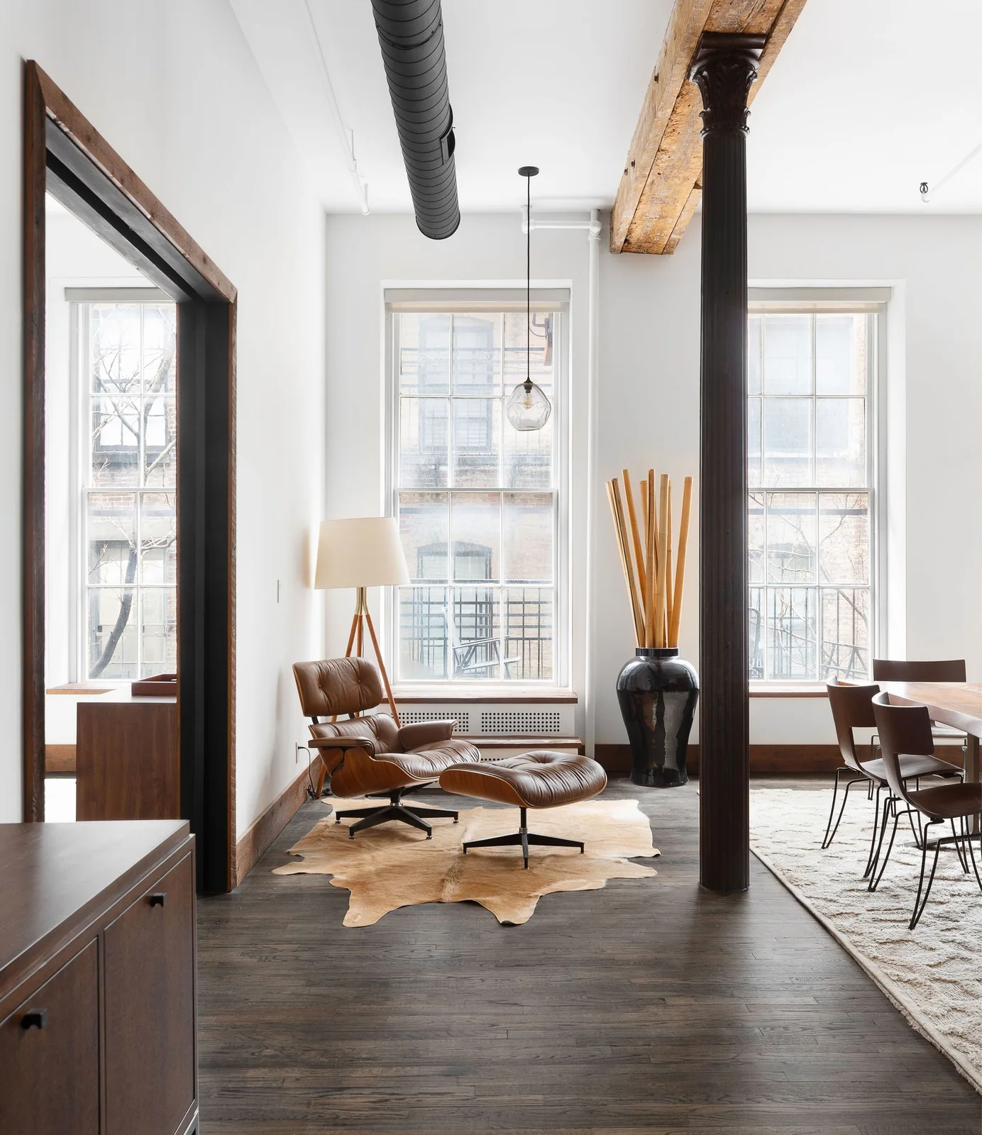 A brown lounge chair and matching footrest sit beside tall loft windows, a floor lamp and an exposed column near a dining area