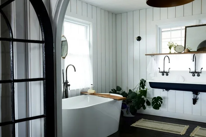 A white freestanding bath and wall-mounted trough basin with dark taps sit beside white vertical boards, a wood shelf and a dark-framed doorway