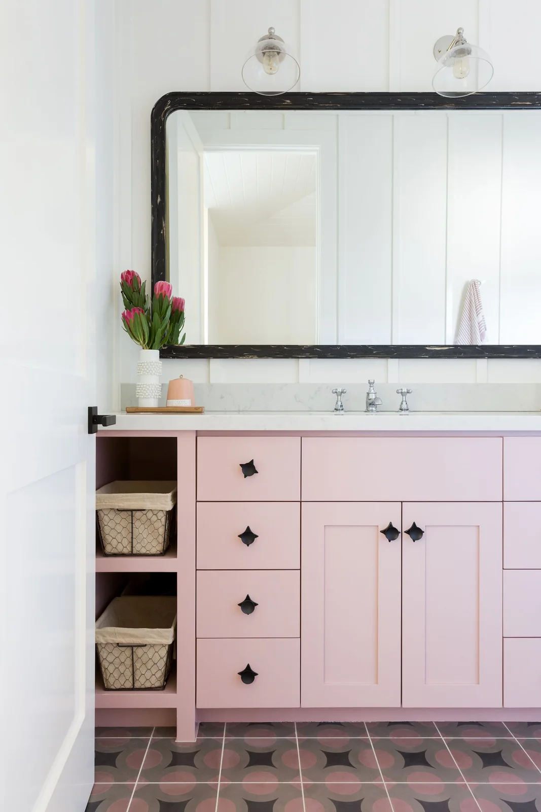 A pink framed vanity with dark knobs and open basket cubbies stands under a broad dark-framed mirror beside white vertical wall boards and pink-and-dark patterned flooring