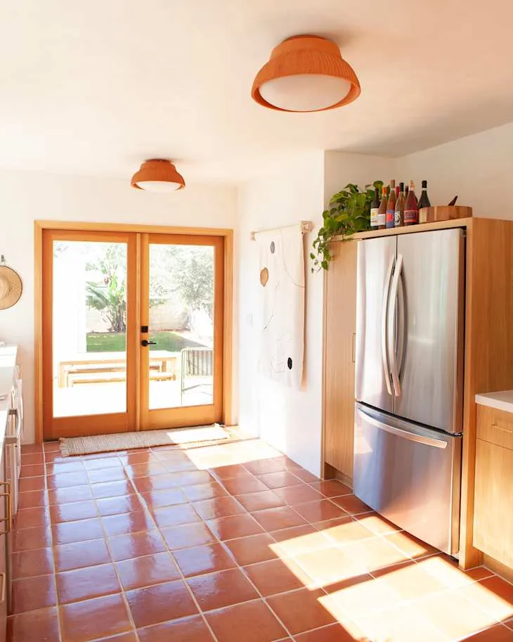 Warm terracotta floor, orange ceiling lights and glass doors opening out from an oak-trimmed kitchen