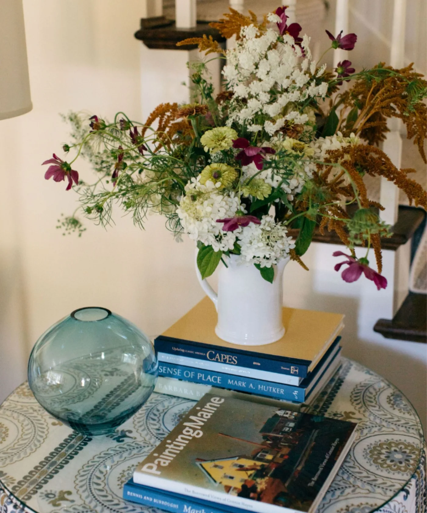 Entry table detail with blue and white patterned cloth, stacked books, blue-green glass bowl and white flower vase.