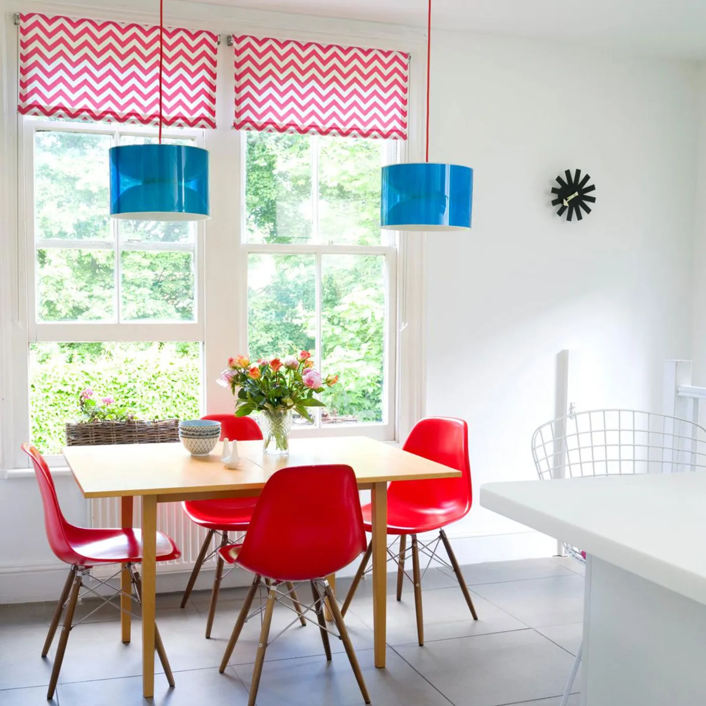 Two blue drum pendants over a rectangular wood table with red chairs and red zigzag window blinds
