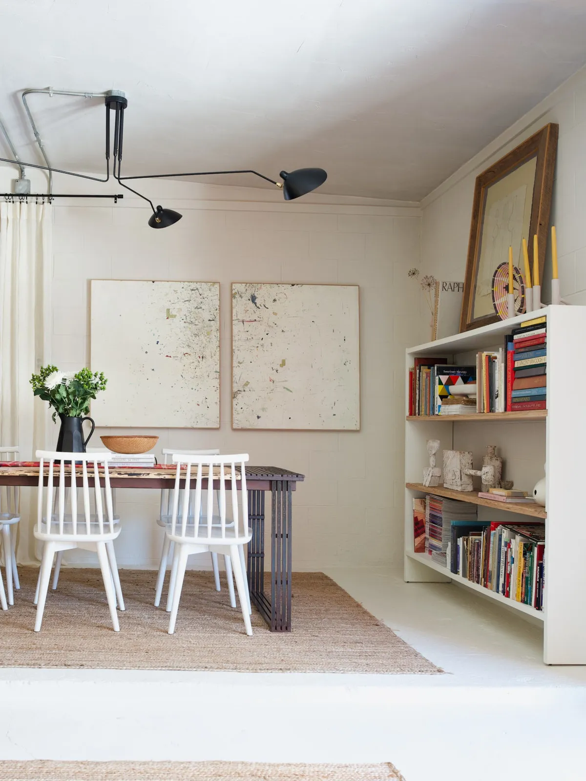 White spindle chairs surround a dark-framed table beside pale artwork, bookshelves and a woven-looking rug