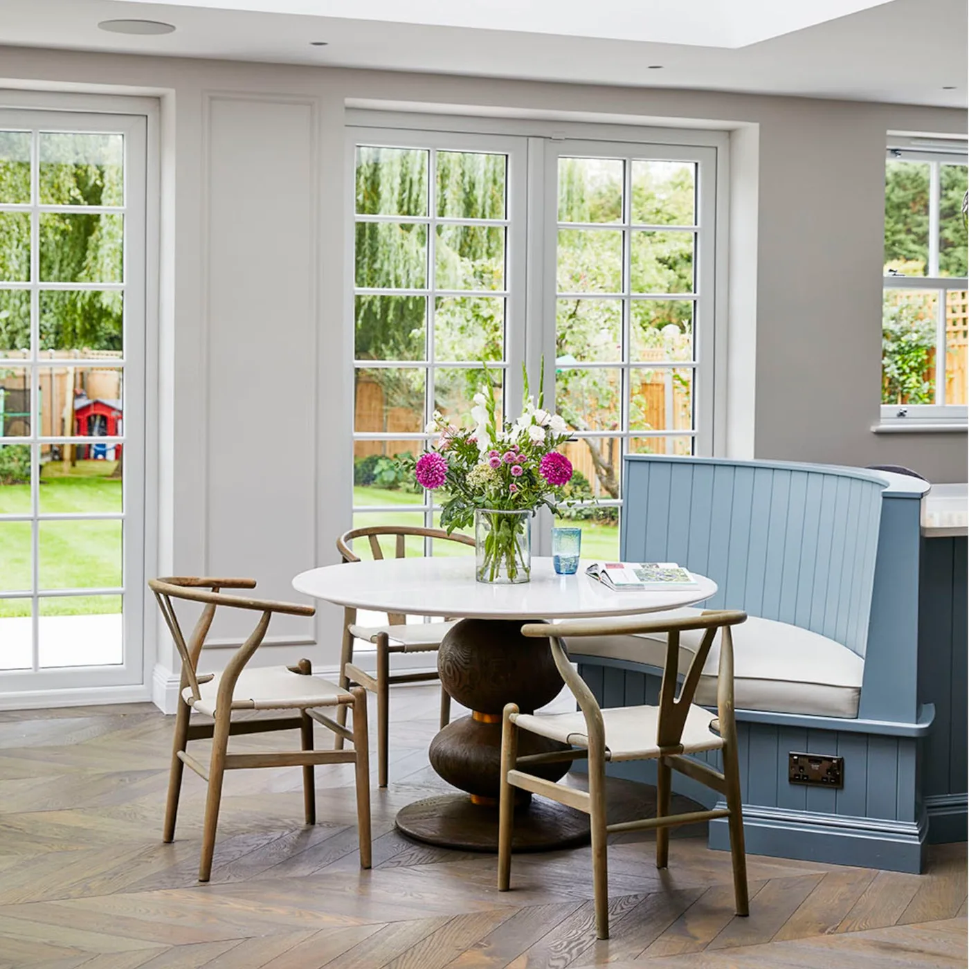 A pale round table with a sculptural dark base sits beside a blue curved bench and wooden woven-seat chairs near broad glazed doors