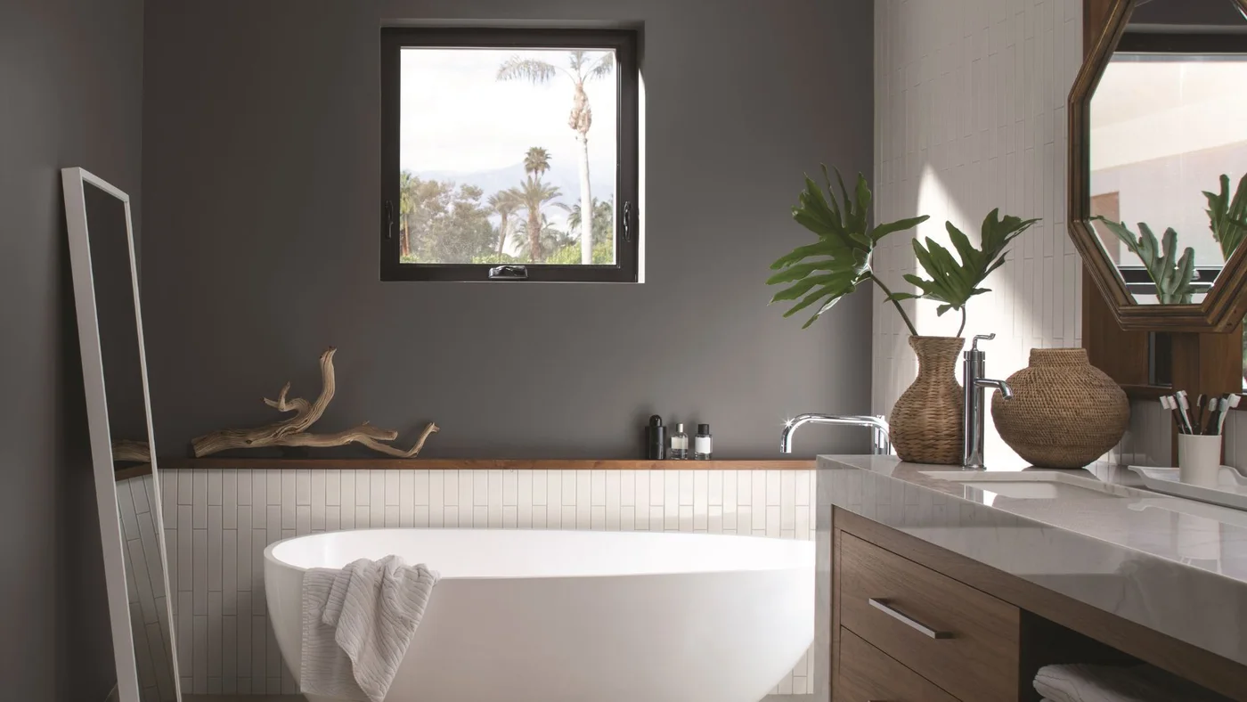 A freestanding white bath sits beside gray walls, a dark-framed window, wooden cabinetry and a large mirror reflecting leafy plants