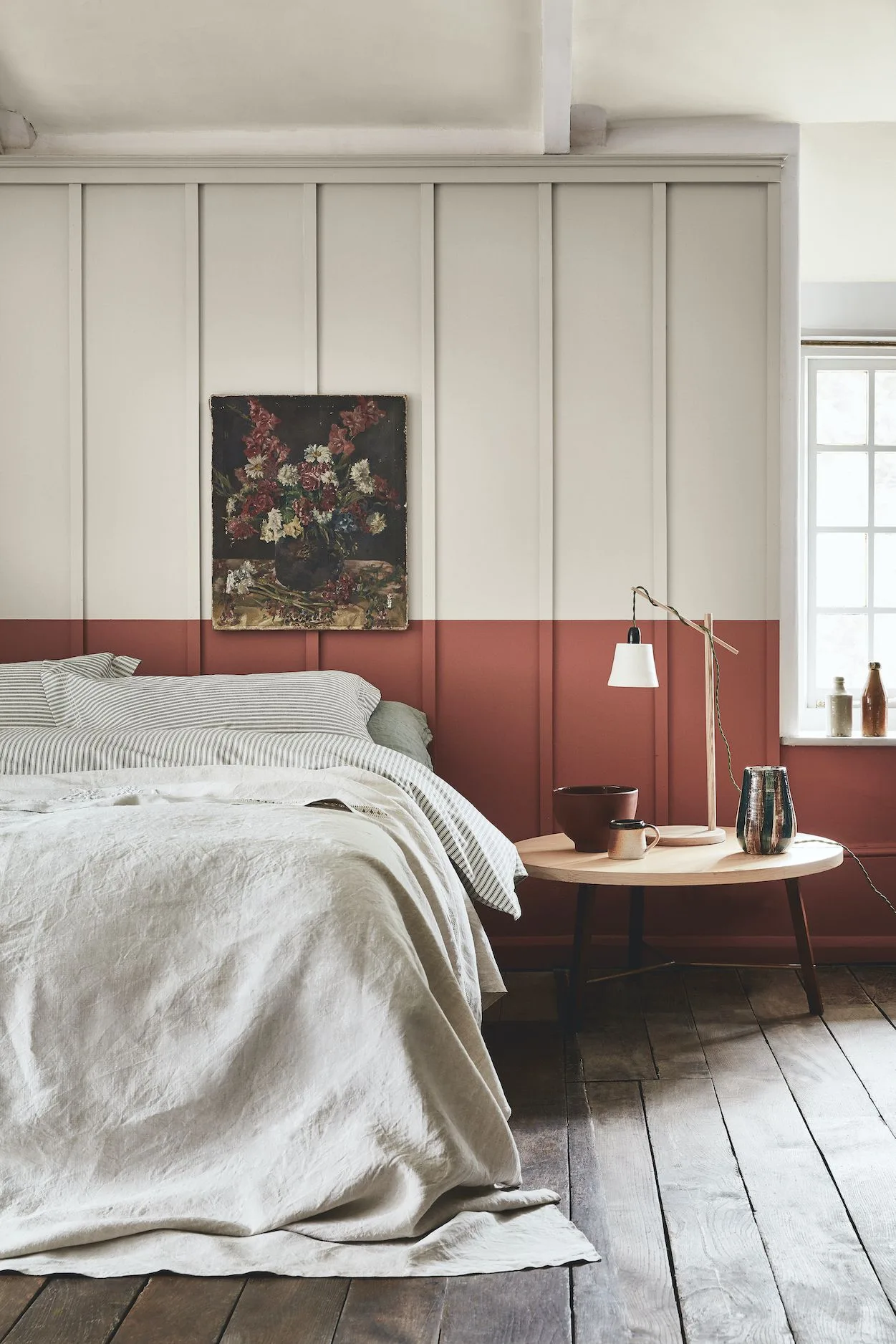 Country bedroom with tall pale wall paneling painted red along the lower section, light bedding, a round wooden side table and a floral picture.