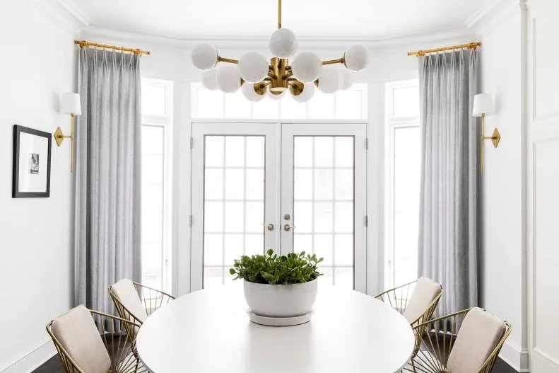 Gold-toned chandelier with white globes above a pale oval table and slender framed chairs, in a bright room with gray curtains and matching wall lights.