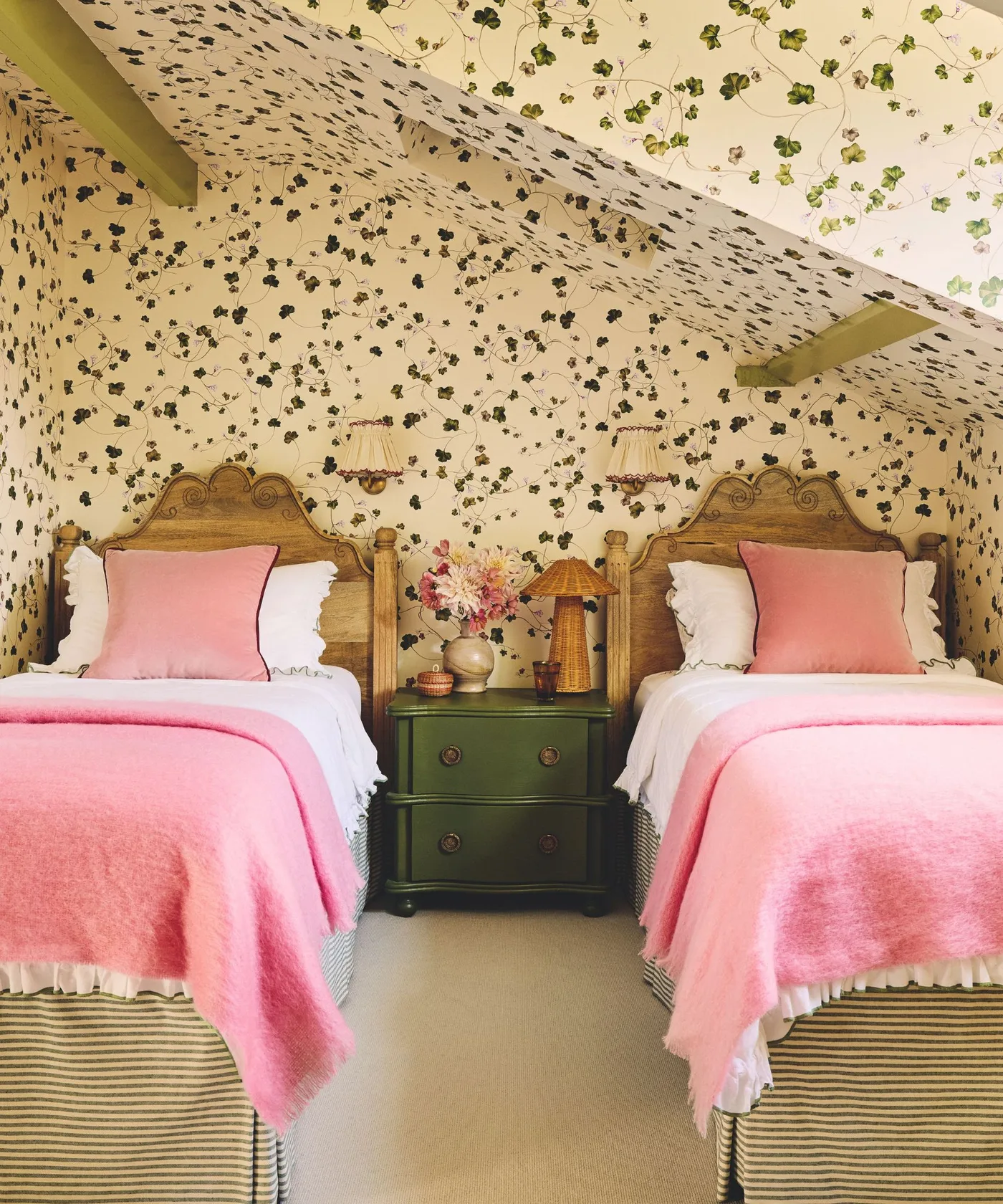 Trailing green ivy wallpaper on attic walls and slopes above twin wooden beds with pink covers and green ceiling beams.