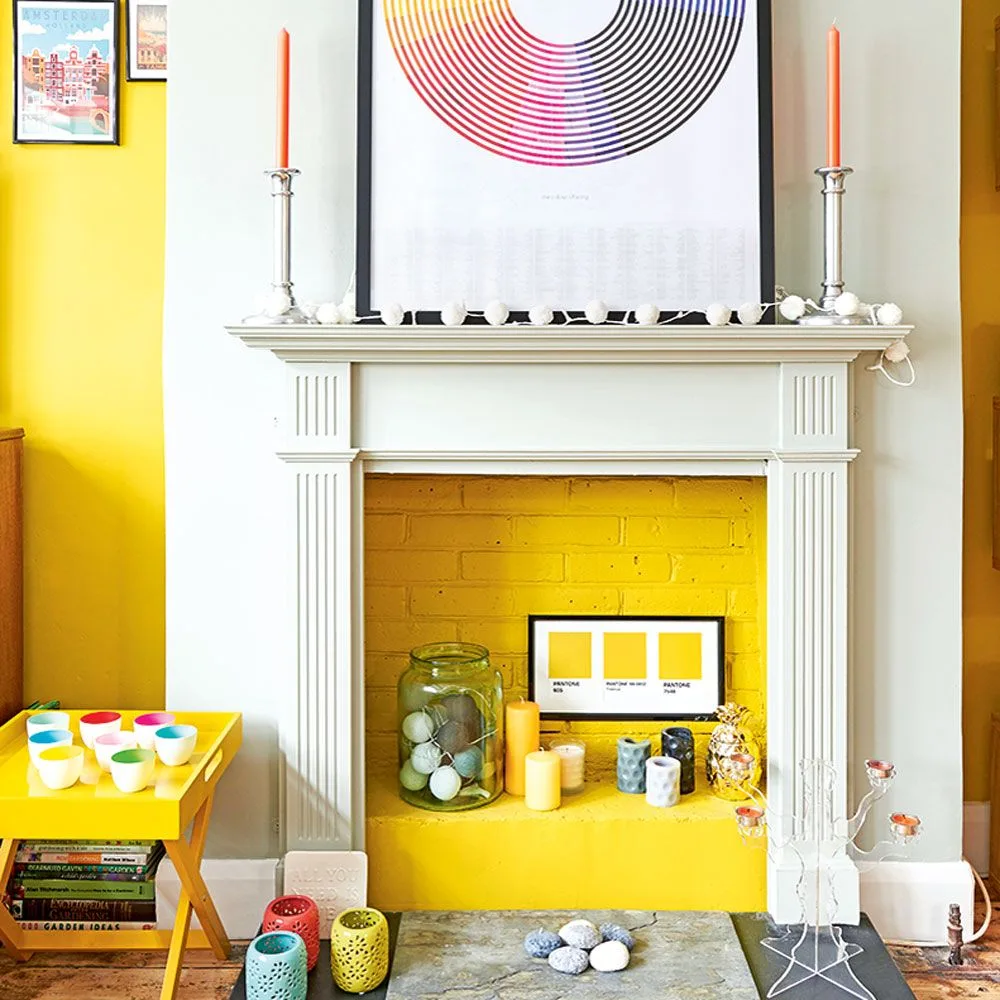 White fireplace with yellow-painted brick opening, colorful pillars below and orange tapers on the mantel.