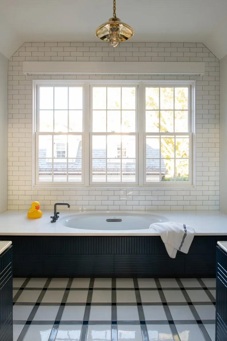 A built-in white bath with a dark paneled front sits below rectangular white wall tiles and a window beside pale checked flooring and a small hanging light