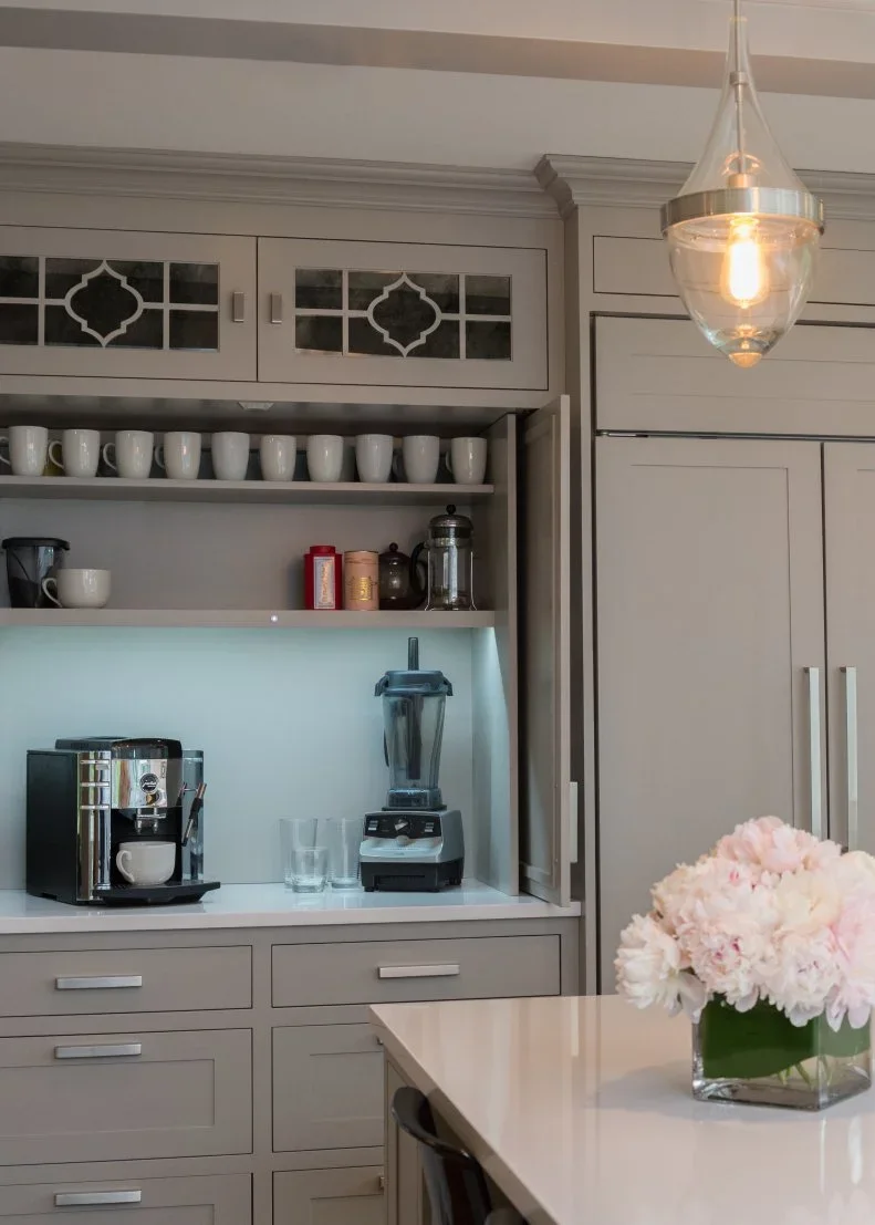 Gray framed cupboards surround a coffee machine and blender beneath an open shelf of white mugs and small glass-front upper doors with straight drawer handles