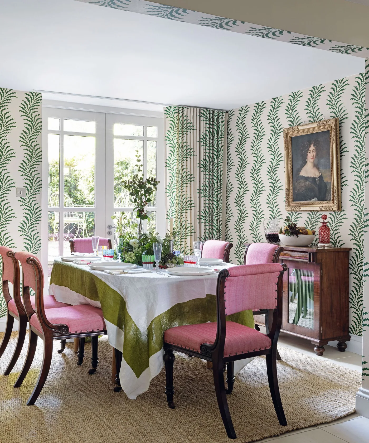 Pink upholstered chairs surround a dressed dining table beside green botanical wallpaper, matching curtains and wooden storage