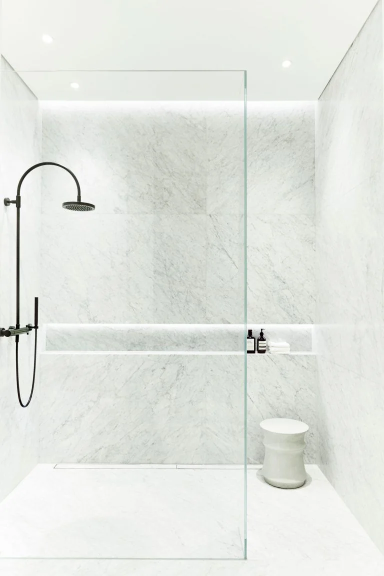 A pale veined shower wall has a black wall-mounted shower fitting above a long illuminated recess beside a glass screen and white stool