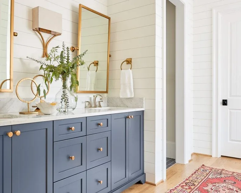 A blue framed bathroom vanity with warm-toned knobs stands beside white horizontal wall boards, tall rectangular mirrors and shaded wall lights