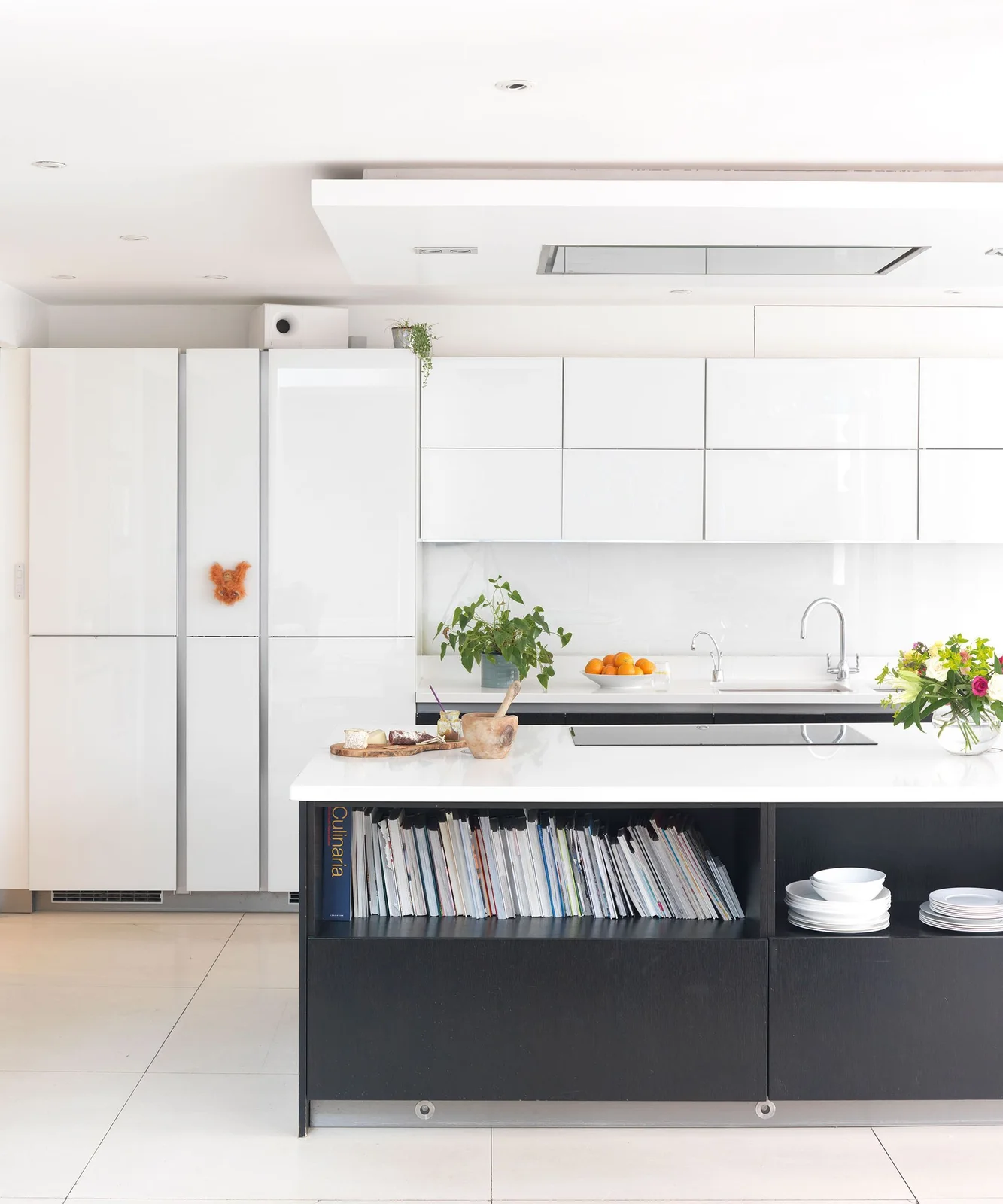 A black kitchen island contains cookbooks and bowls below a white counter beside white cabinets