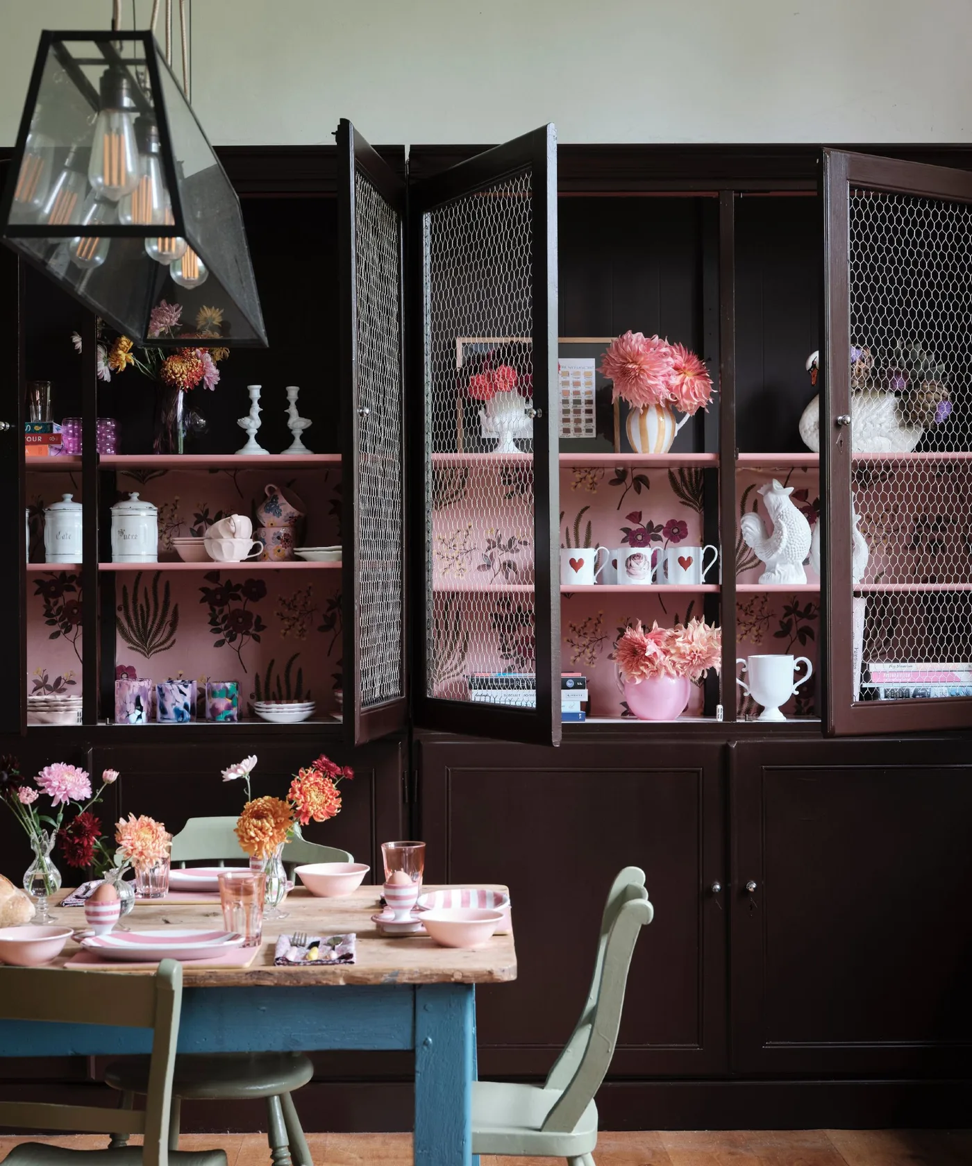 Dark country cupboard with pink floral paper behind shelves of cups and serving pieces