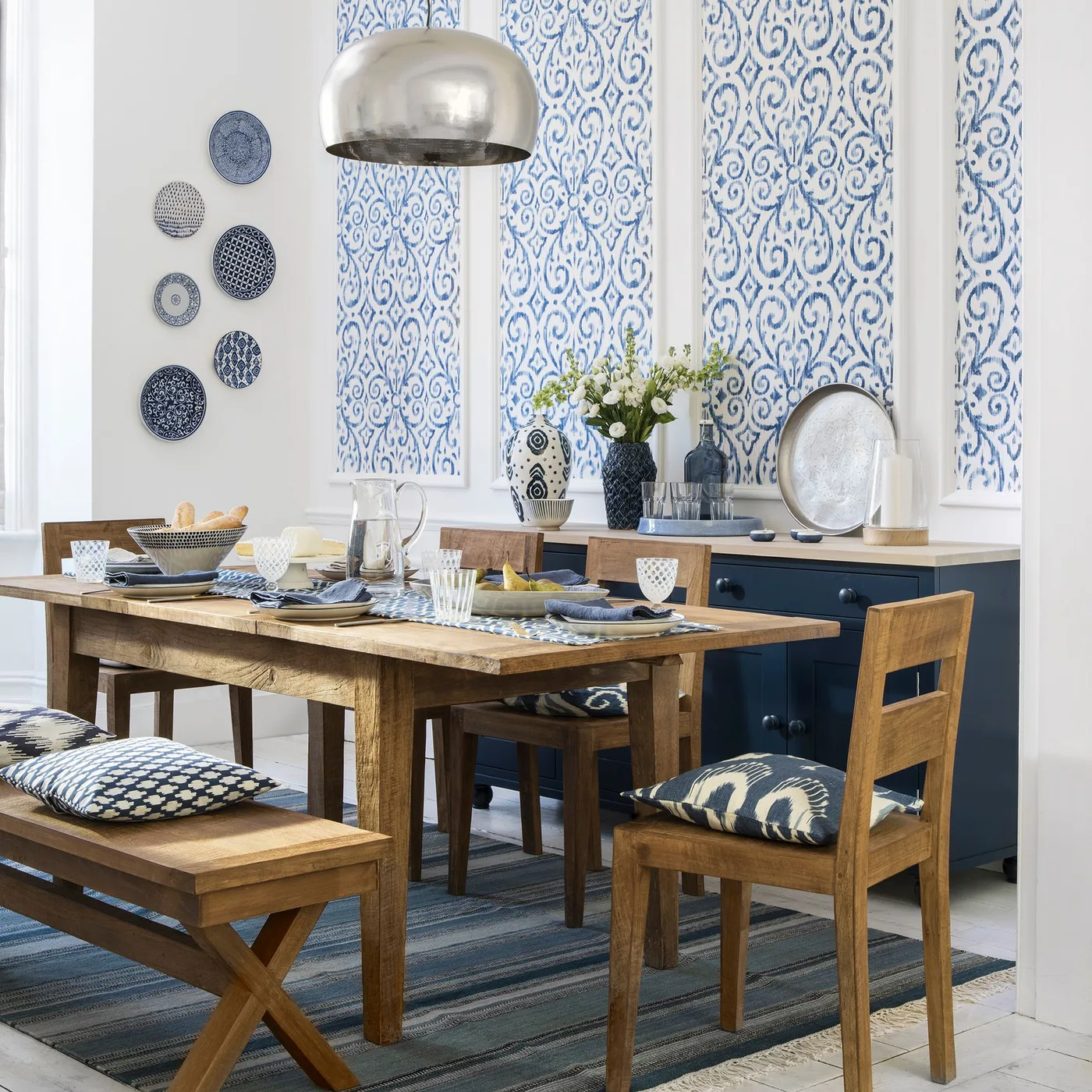 Cottage dining room with blue patterned wallpaper inside pale wall frames, wooden table and bench, blue sideboard and patterned cushions.