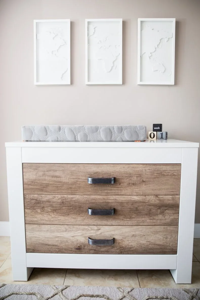 White changing-area unit with wood-toned drawer fronts beneath three pale framed pictures.