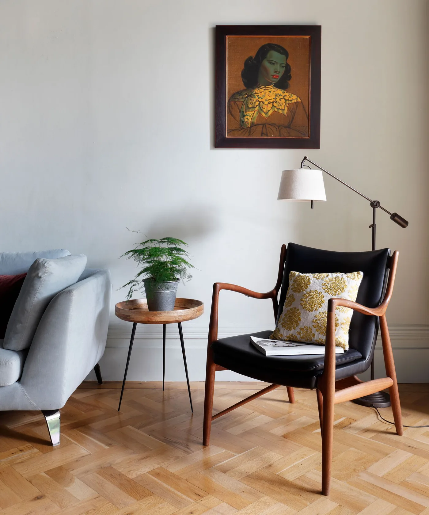 A wood-framed dark reading chair stands beside a round wood side table and adjustable lamp against a pale wall above a warm parquet floor