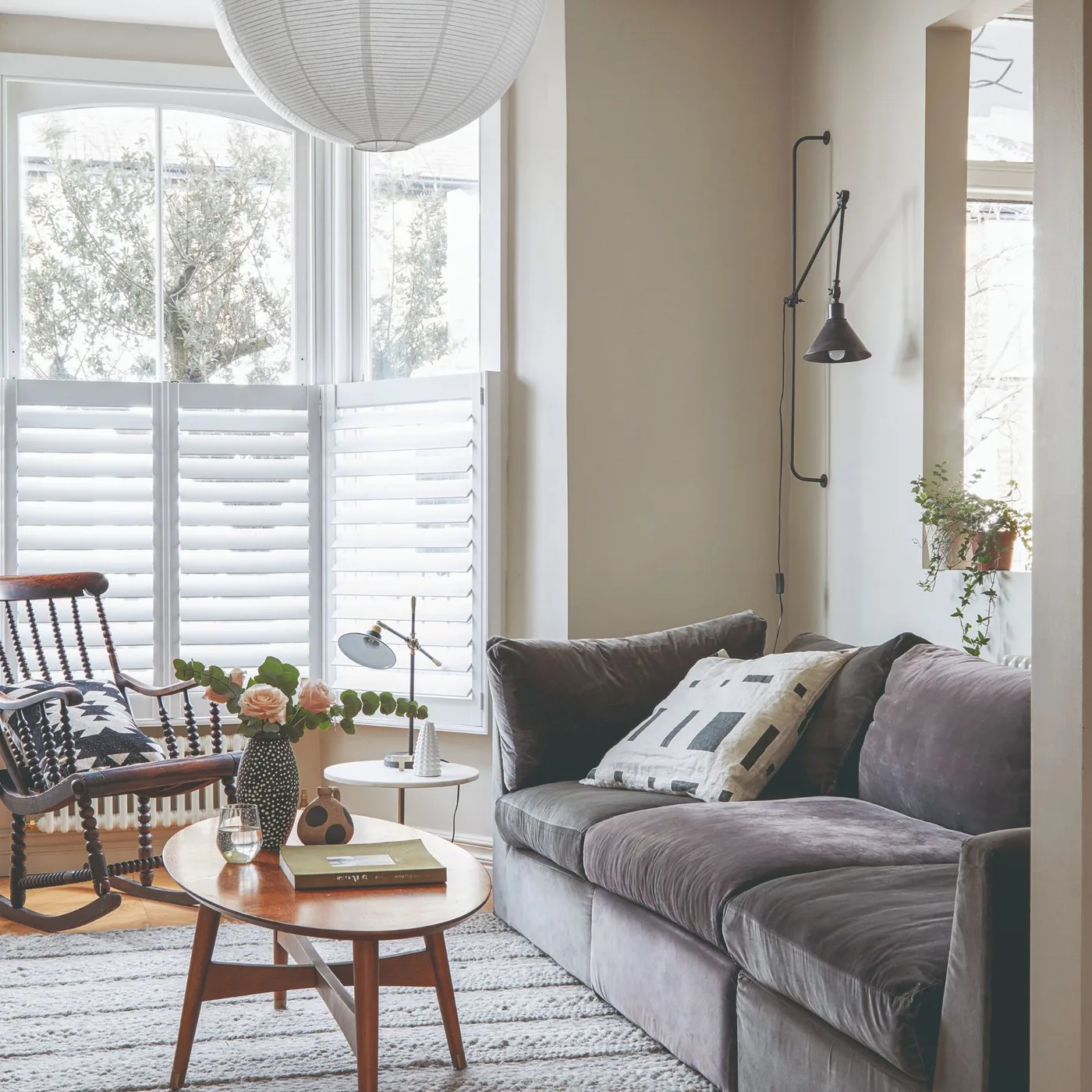 A gray sofa sits beside a round wood table, a wood rocking chair, pale window shutters and a slim adjustable wall lamp