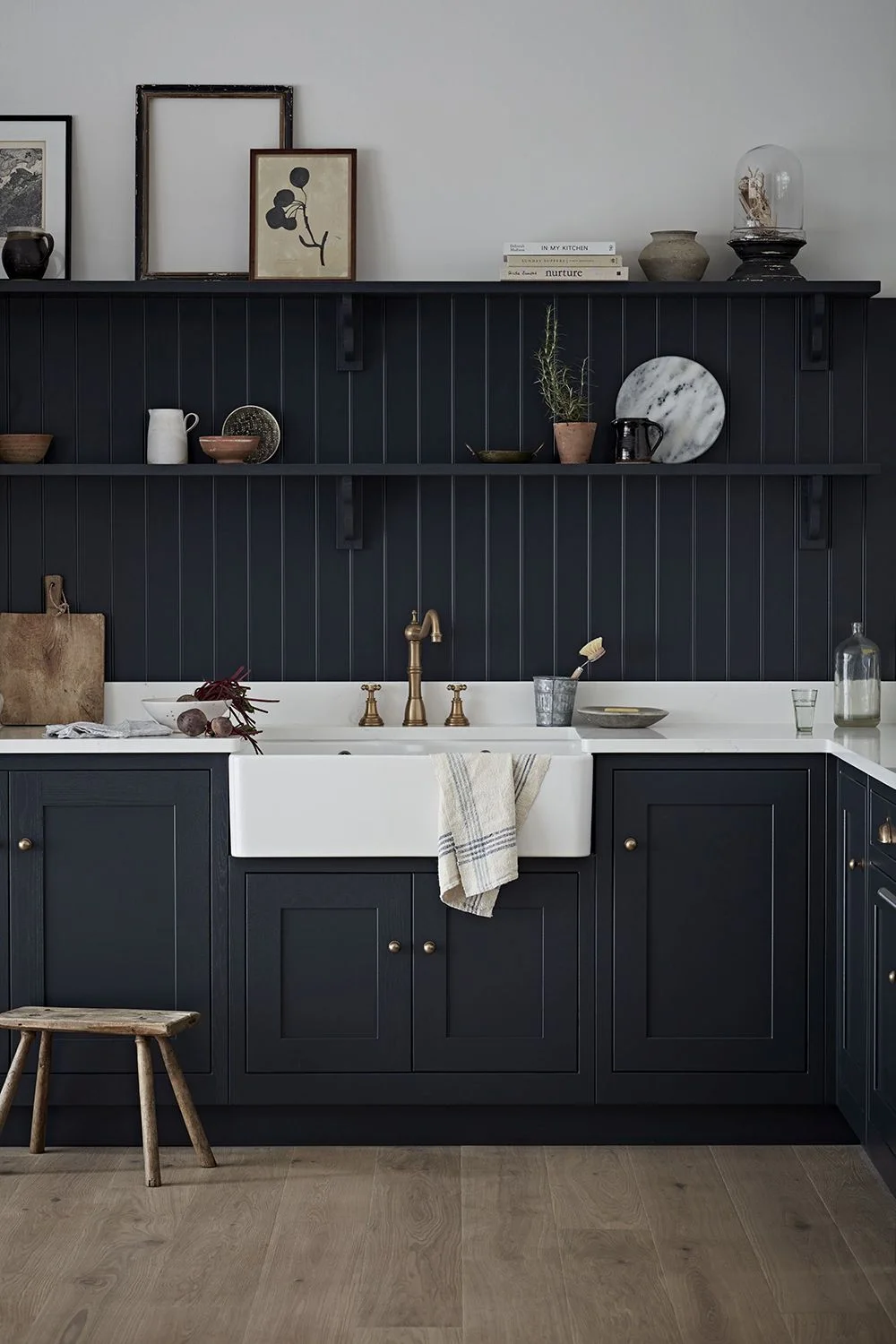 A white apron-front kitchen sink sits among dark framed cabinets below dark vertical wall paneling and shelves beside lighter upper walls