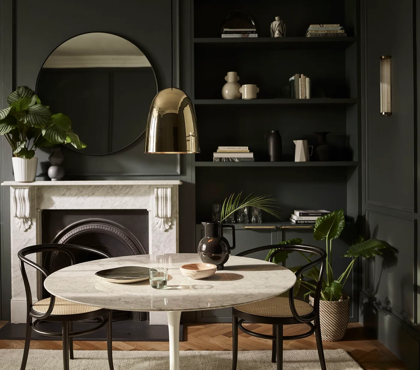 A round pale dining table and dark open-backed chairs sit beside deep green shelving, a pale fireplace surround, round mirror and warm-colored pendant