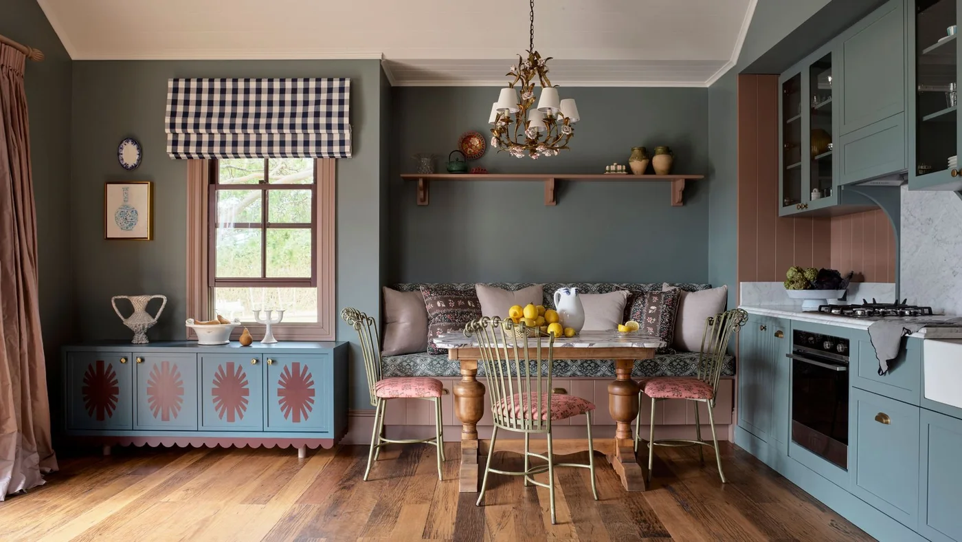 Blue-gray cabinets and a painted decorative sideboard sit beside a checked blind, patterned bench cushions, pink chair seats and a decorative chandelier