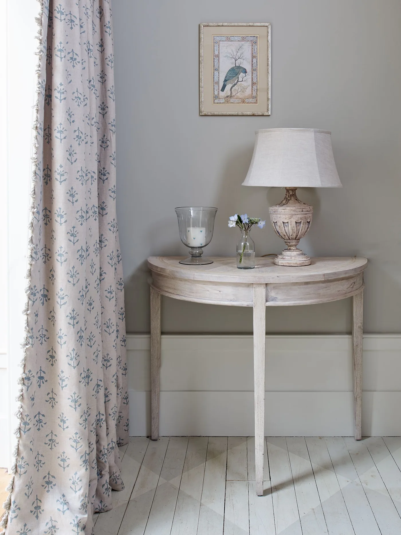Country entry detail with a pale half-round console table, small lamp, bird picture and a light patterned curtain against a pale wall.