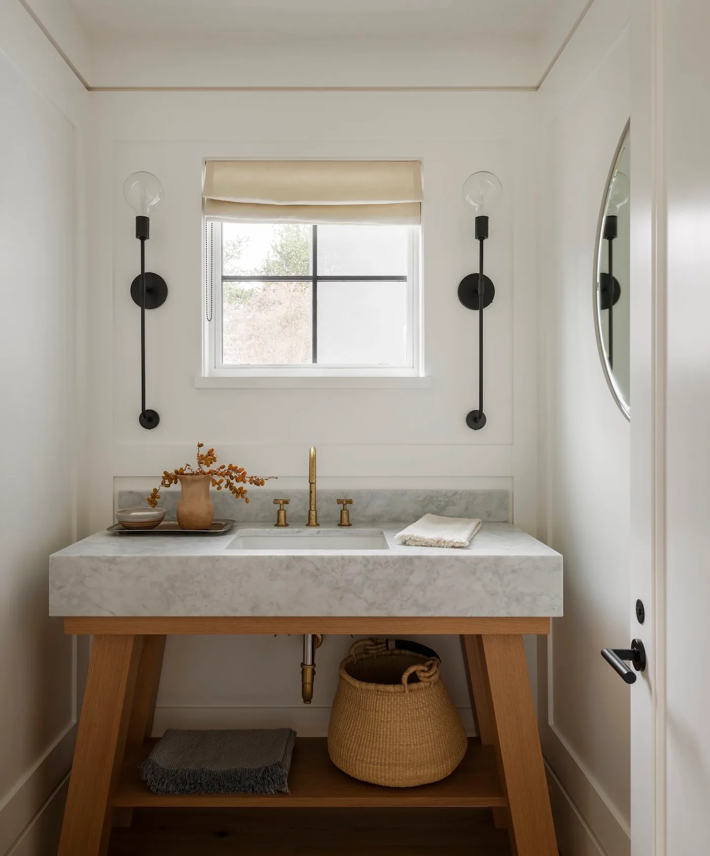 Powder room with pale basin counter on wooden frame, round mirror, black wall lights, small window, pale shade and large woven basket on lower shelf.
