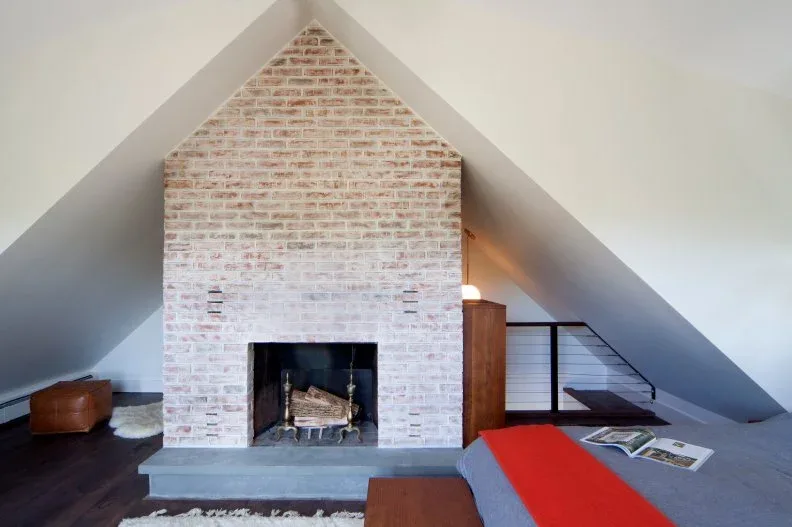 Pale mottled brick fireplace following a steep attic roofline beside a bed with a red throw.