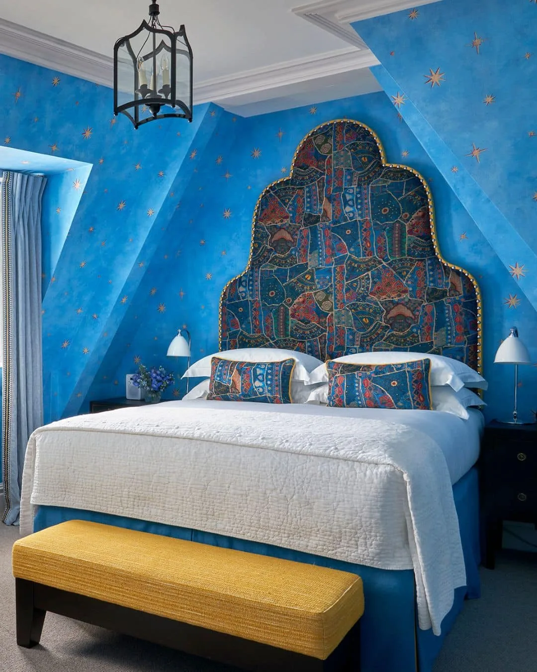 Blue star-patterned sloping bedroom walls around a tall patterned headboard, white bedding and a yellow bench.