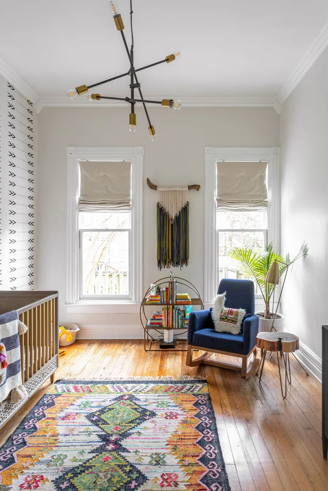 A multicolored geometric rug lies beside a navy adult rocker, round book stand and patterned crib wall beneath a branching ceiling light