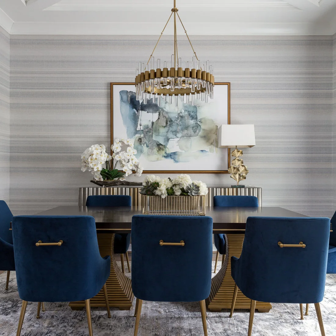 A ridged metallic-looking sideboard stands beneath blue-toned artwork and a pale lamp behind blue upholstered dining chairs
