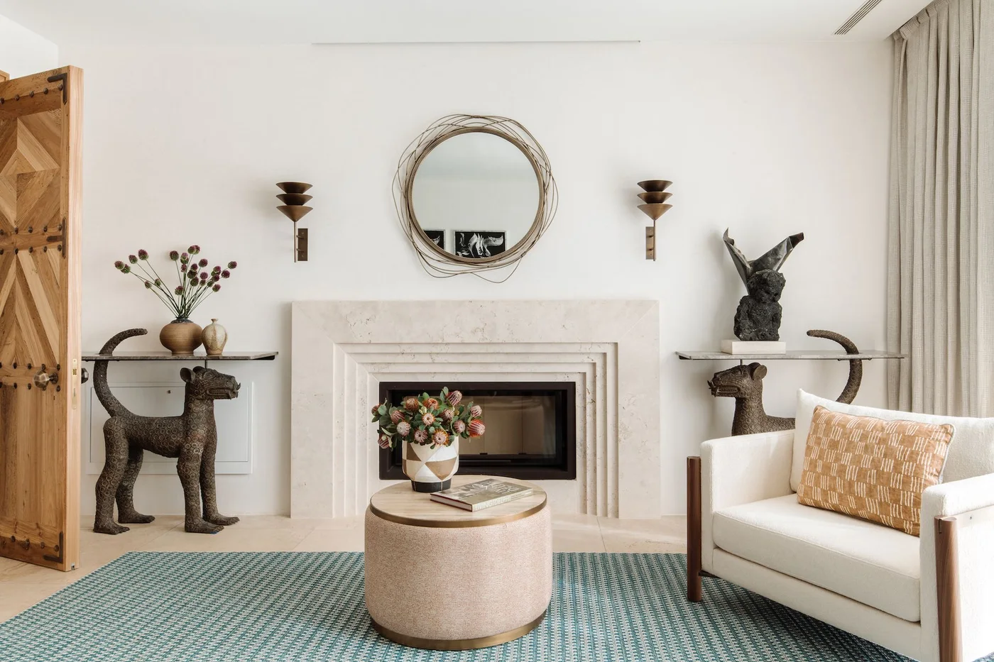 A pale stepped fireplace frame sits under a round mirror between dark wall lights beside a white chair, round table and green patterned rug