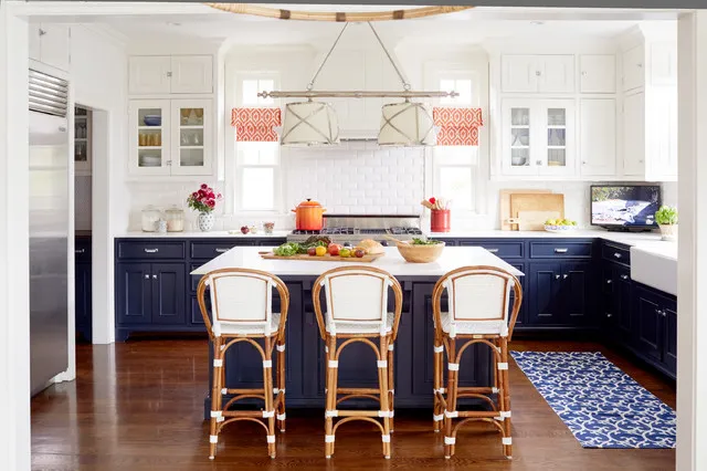 Navy lower cabinets and an island stand below white upper cupboards beside woven-backed stools, patterned red window blinds and a blue patterned runner