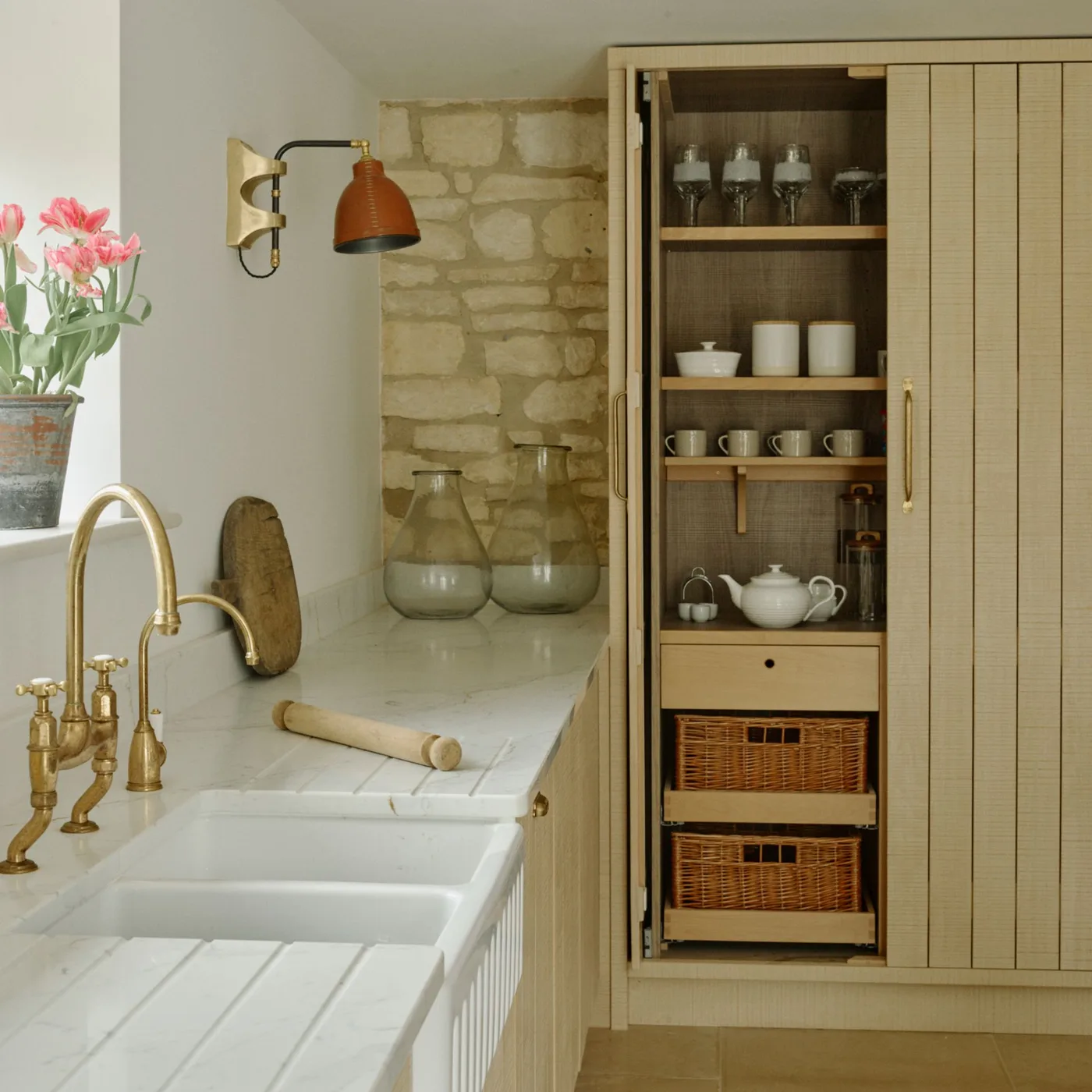 Pale kitchen with a white apron-front sink, gold-colored tap, open tall pantry and baskets in its lower section.