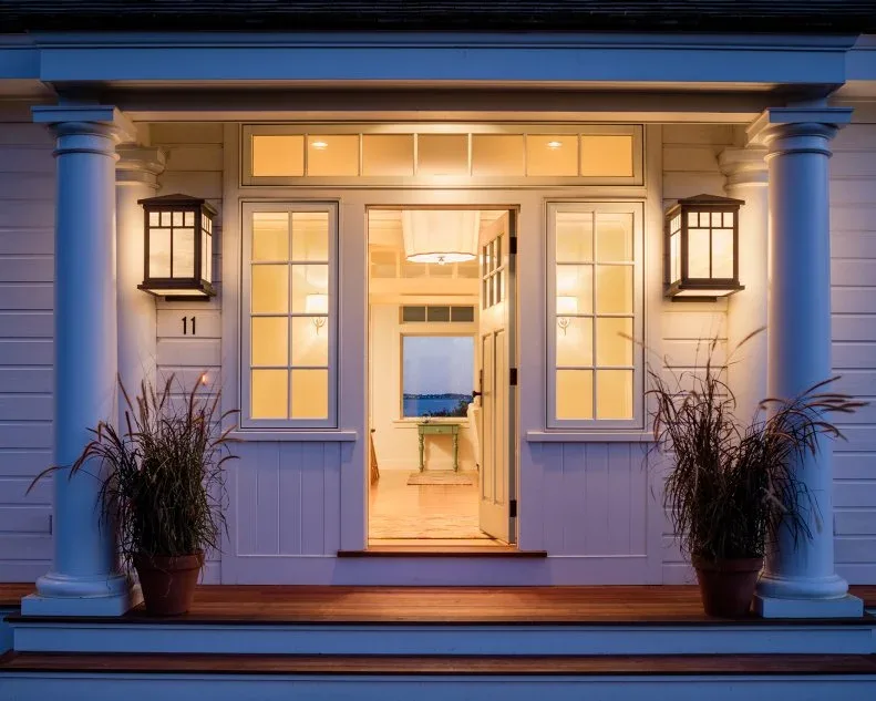 Pale cottage entrance at dusk with glazed door, sidelights, transom, paired lanterns and tall planted pots