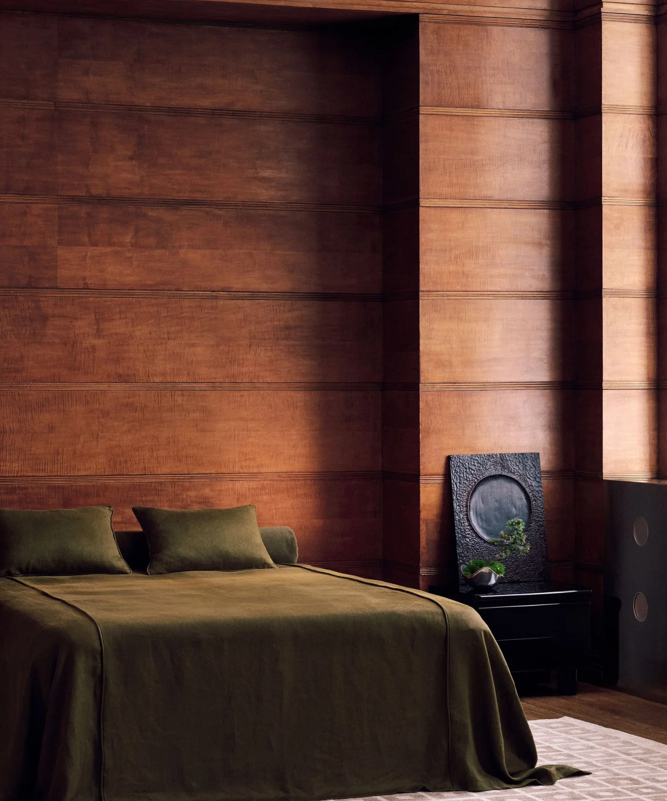 Deep wooden wall panels surround a bed with olive-green bedding beside a dark bedside cabinet and framed artwork