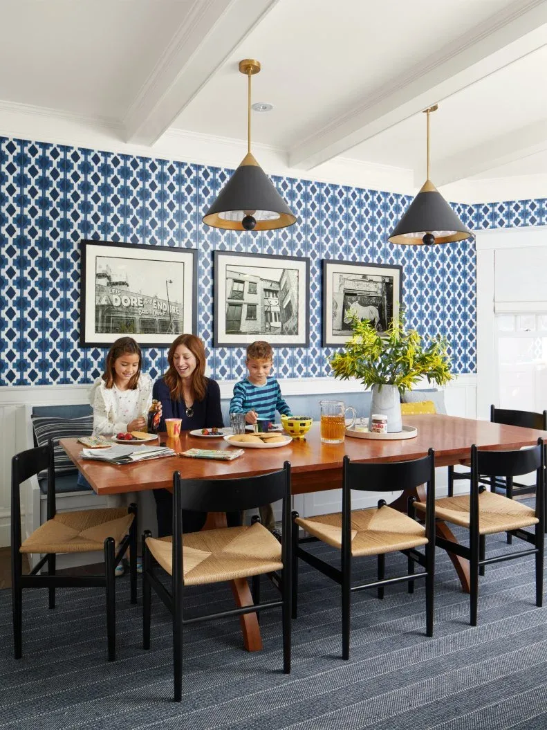 Dining area with blue-and-white geometric wallpaper behind a bench, three framed photographs, wooden table, black chairs with pale seats, and two dark pendants.