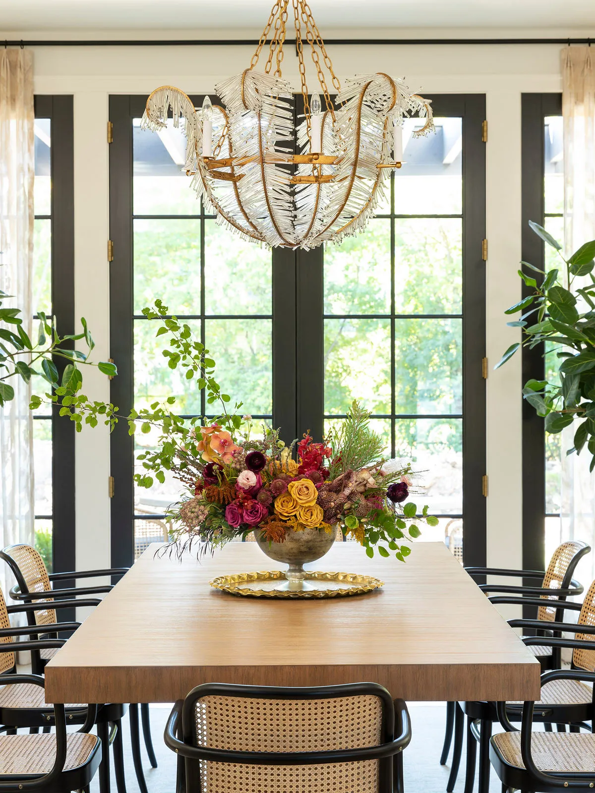 Cane-backed chairs surround a long timber table beneath a decorative chandelier beside large windows.