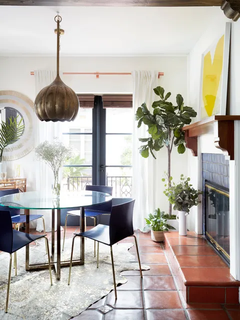 A small dining table sits beneath a textured pendant beside plants and a large wall mirror.