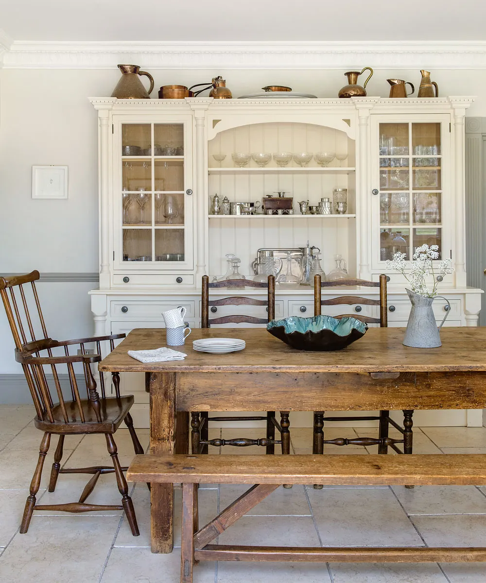 A tall cream dresser with glazed doors stands behind a wooden dining table and bench