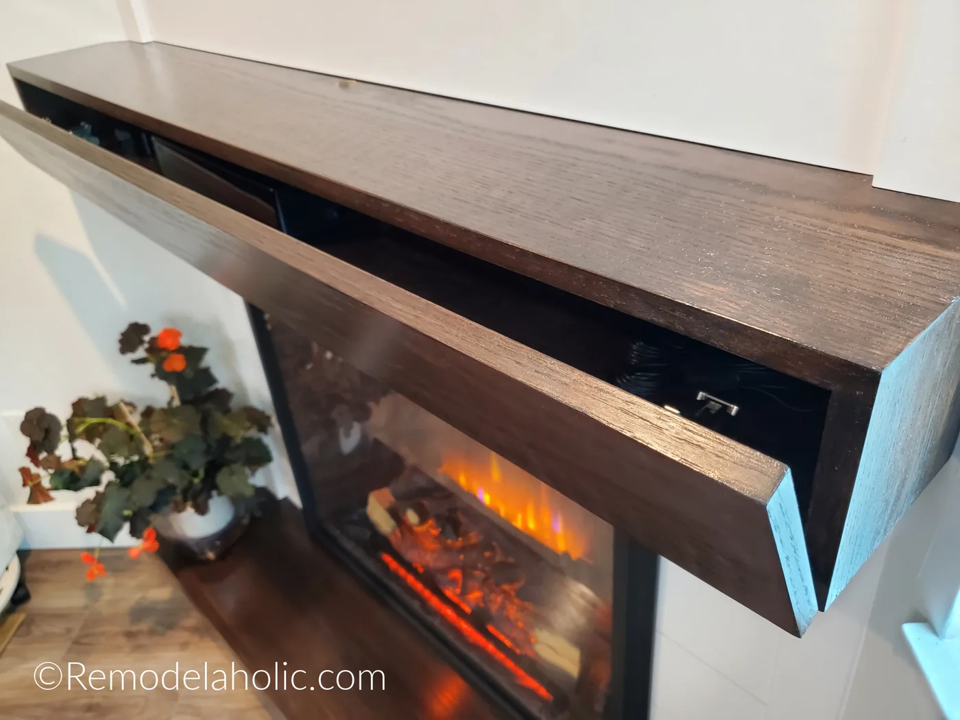 Close view of an open wood mantel compartment above an electric fireplace, with decorative flowers beside it.
