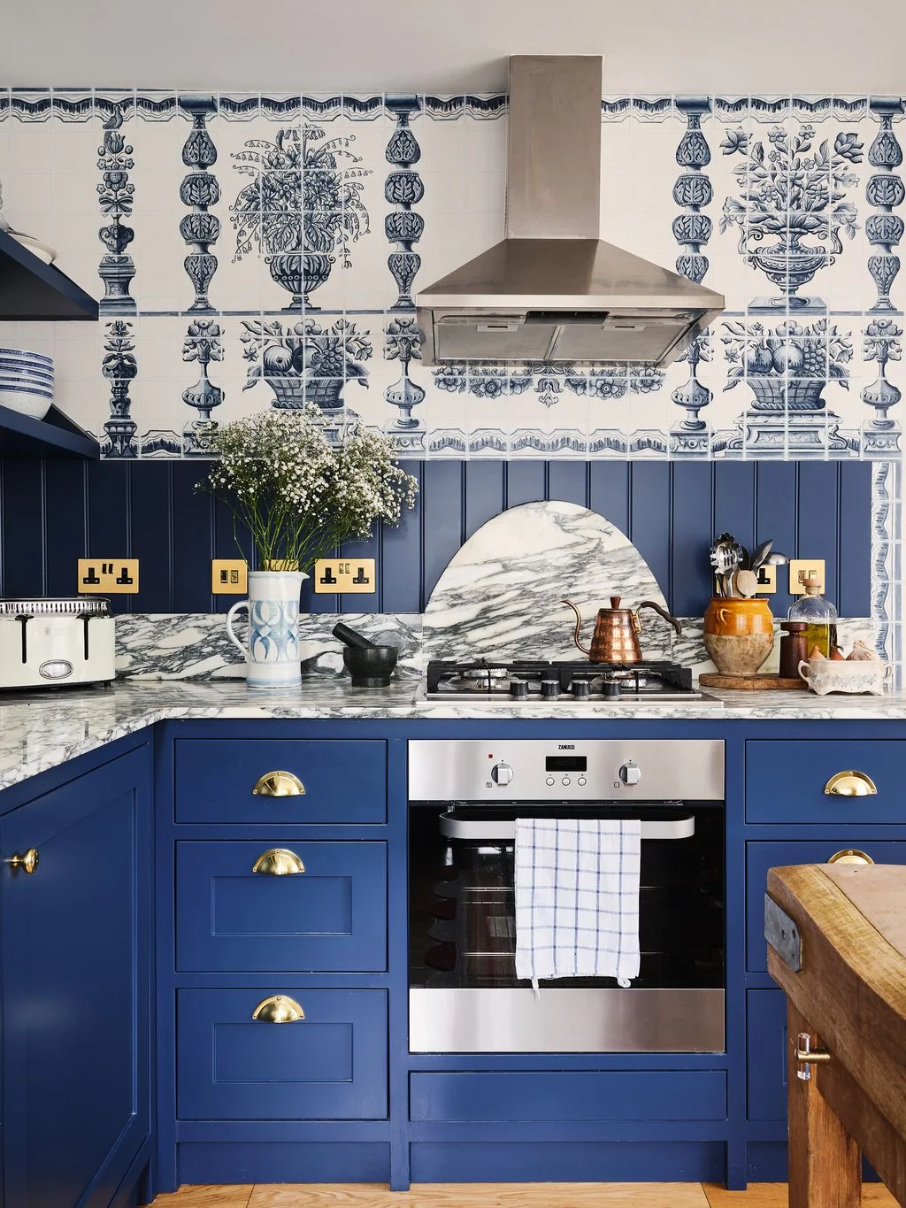 Deep blue cabinets and paneling sit beneath blue-and-white illustrated wallpaper with decorative vase motifs beside a veined countertop and silver-colored hood
