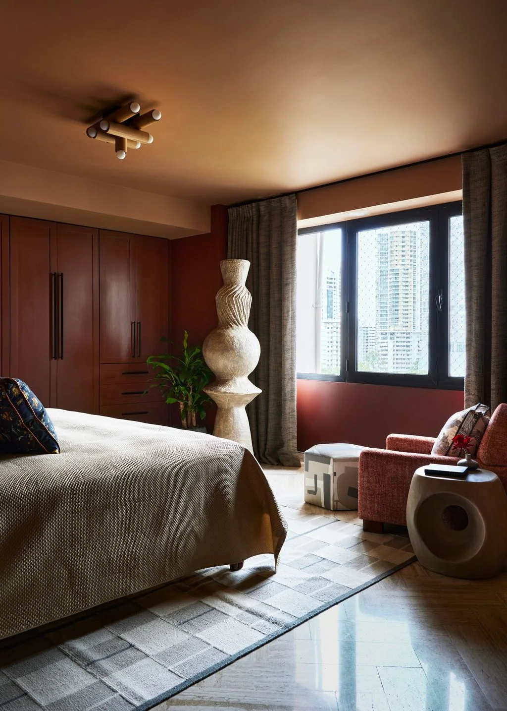 A pale-covered bed stands beside warm reddish wardrobe doors and walls, gray curtains, a reddish chair and sculptural side table near a dark-framed window