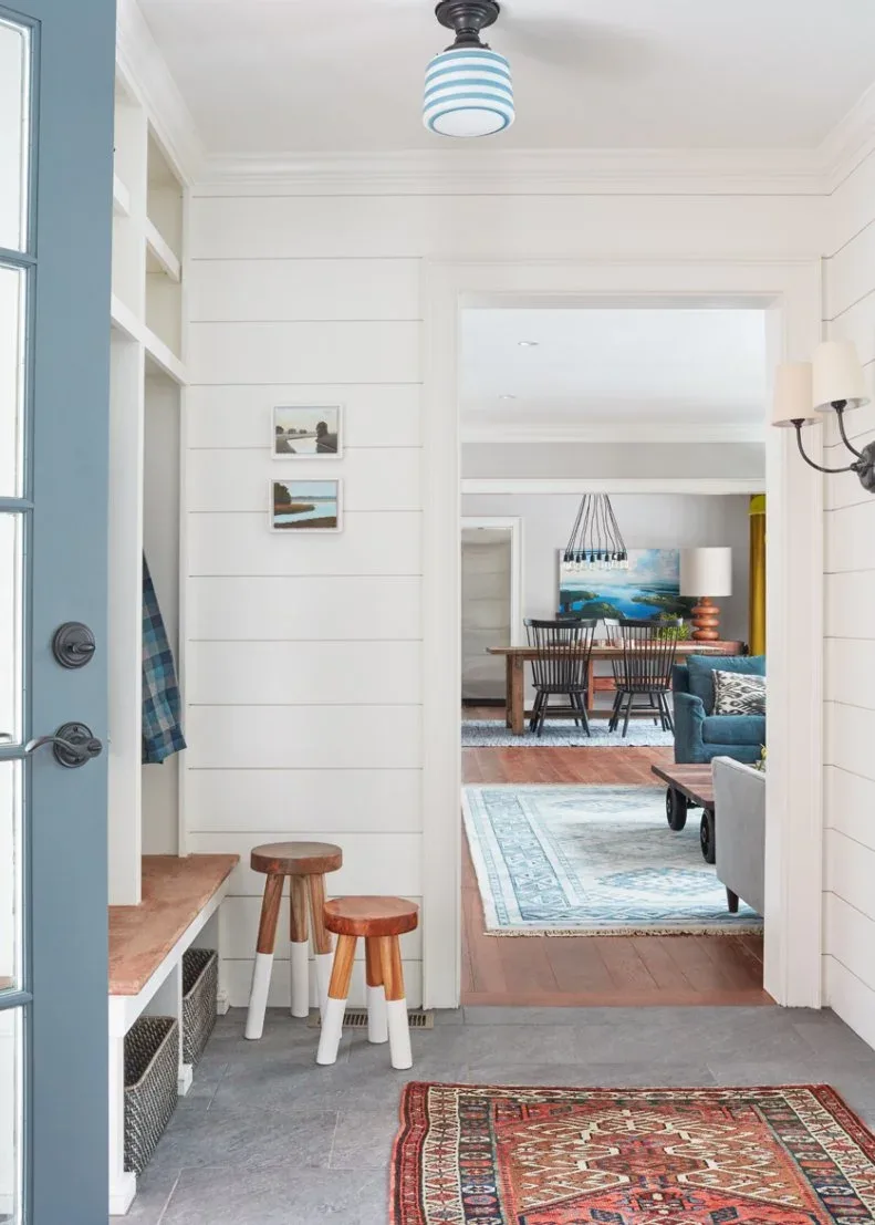 White boarded entry with wooden-topped bench, lower baskets, two small wooden stools and red patterned rug.