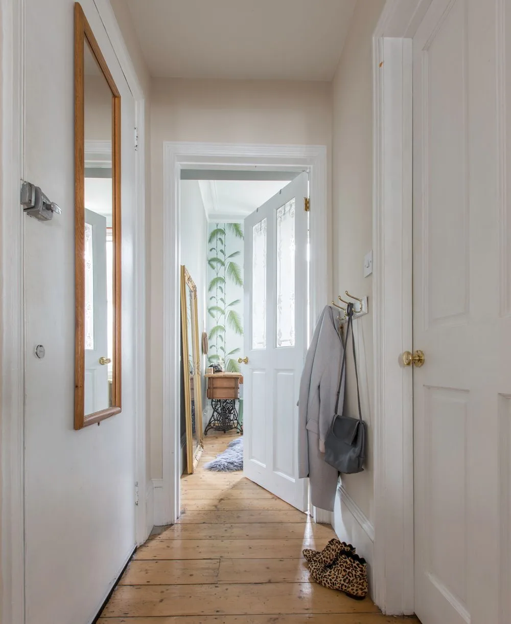 White paneled hallway with a tall wooden-framed mirror, coat hooks, warm floorboards and open internal doors.