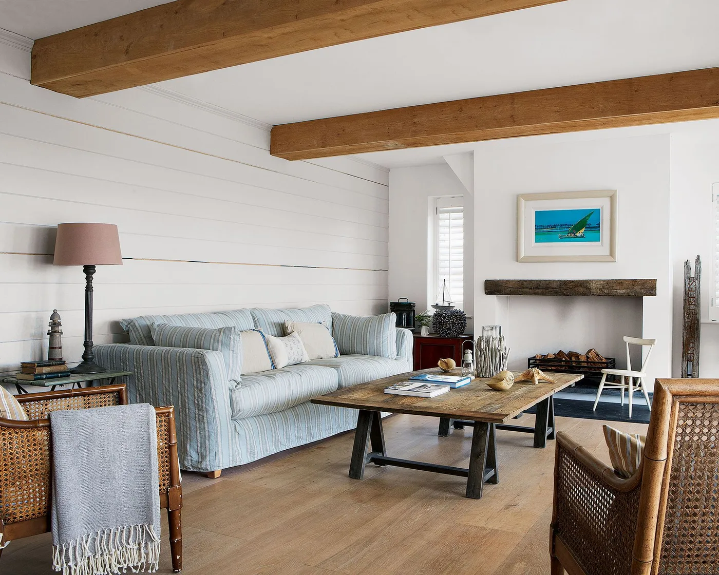 Pale paneled living room with exposed wooden beams, a striped light sofa, wooden coffee table and woven-back chairs.