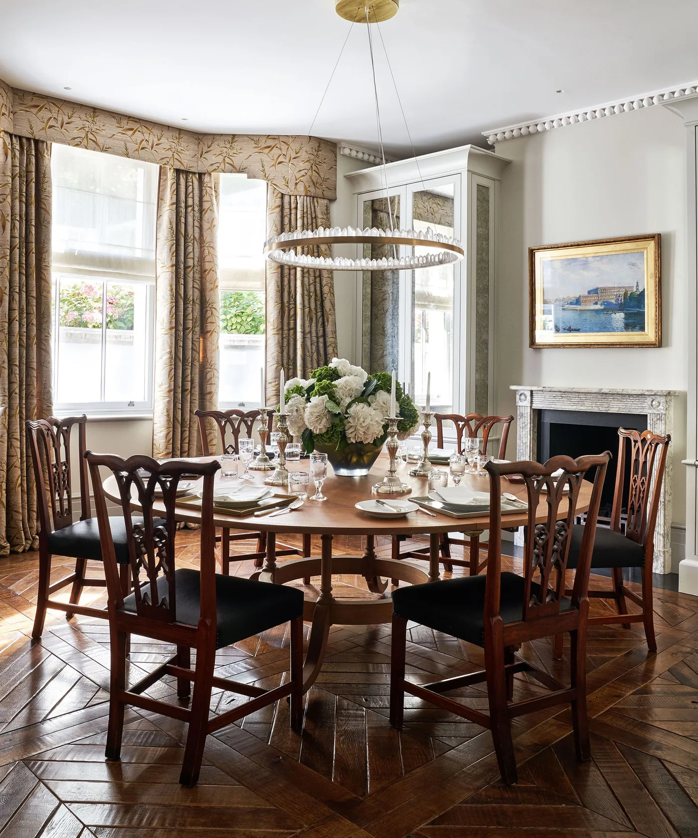 Formal dining room with a round pedestal table, carved wooden chairs, patterned curtains and a pale fireplace surround.